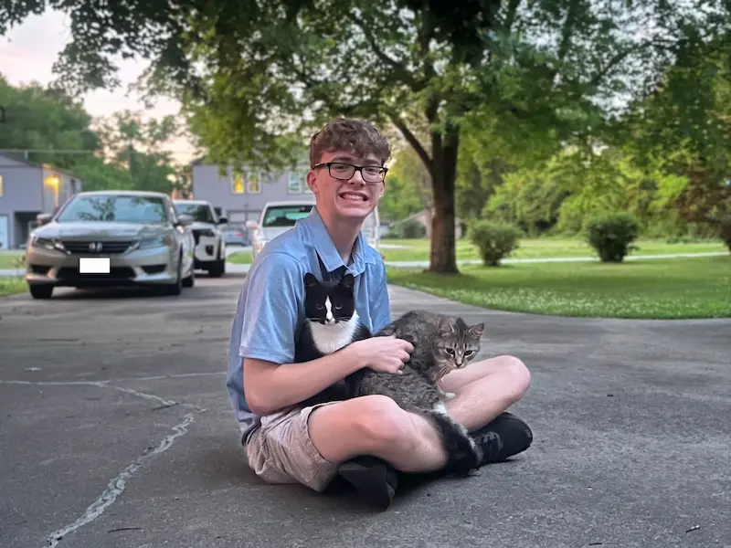 Jake, founder of Fairstrive sitting cross-legged on a driveway holding a tuxedo cat and a tabby cat in his lap.